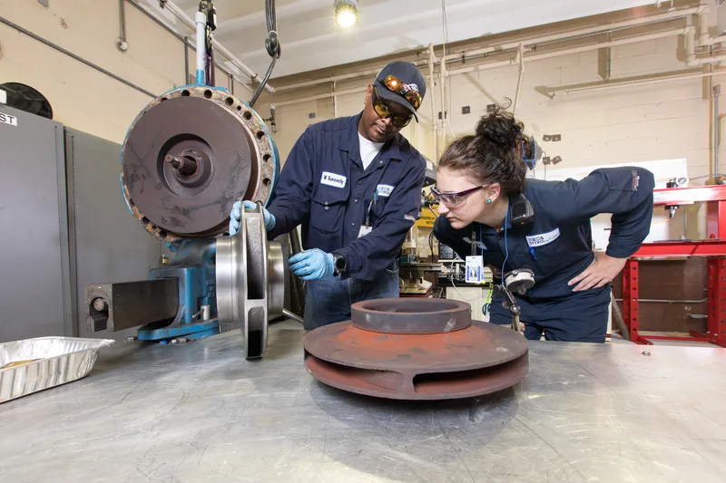 Refurbished equipment technicians handling worn cast iron impeller beside stainless steel machinery