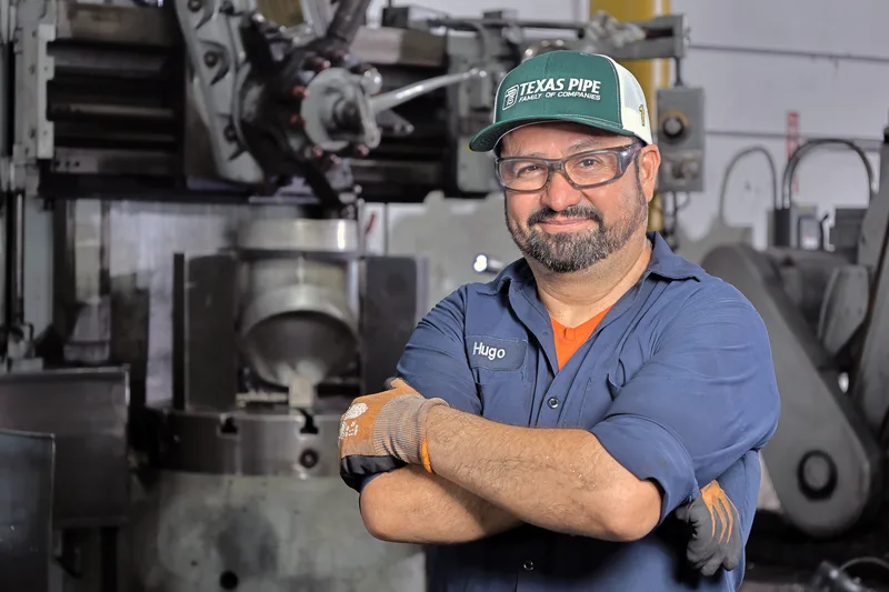 smiling worker in safety glasses and gloves posing arms crossed beside metal lathe