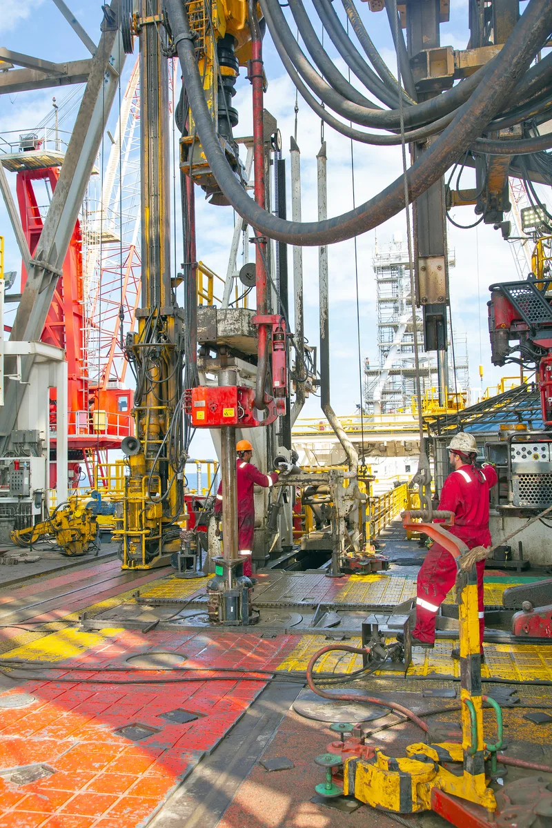 Offshore rig drilling floor workers in red coveralls operating hydraulic pipe equipment