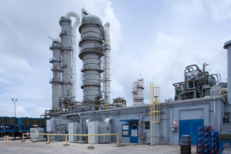 Styrene control room exterior beside towering gray distillation columns and yellow guardrails